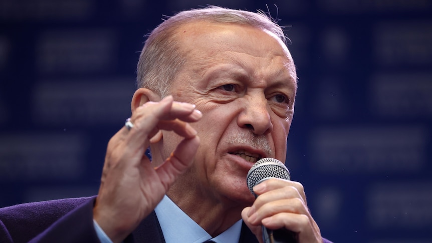 An older Turkish man in a suit yells into a microphone.