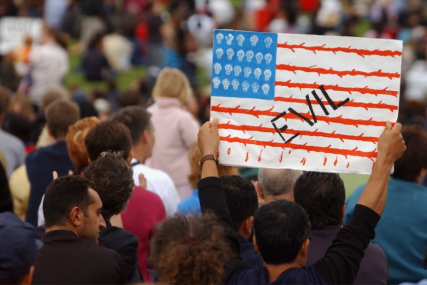 Men stand amid crowd holding a painting of the US flag with skulls insead of stars and "EVIL" brandished in black letters