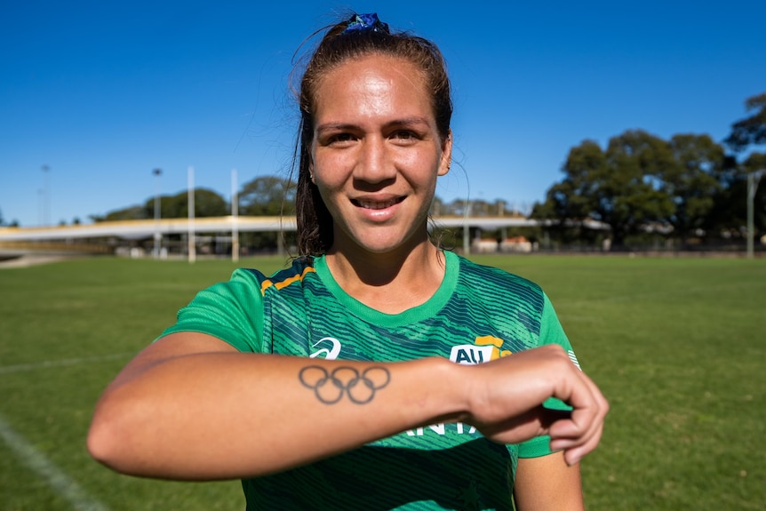 An Australian women's rugby sevens player smiles at the camera and displays an Olympic rings tattoo on her arm.