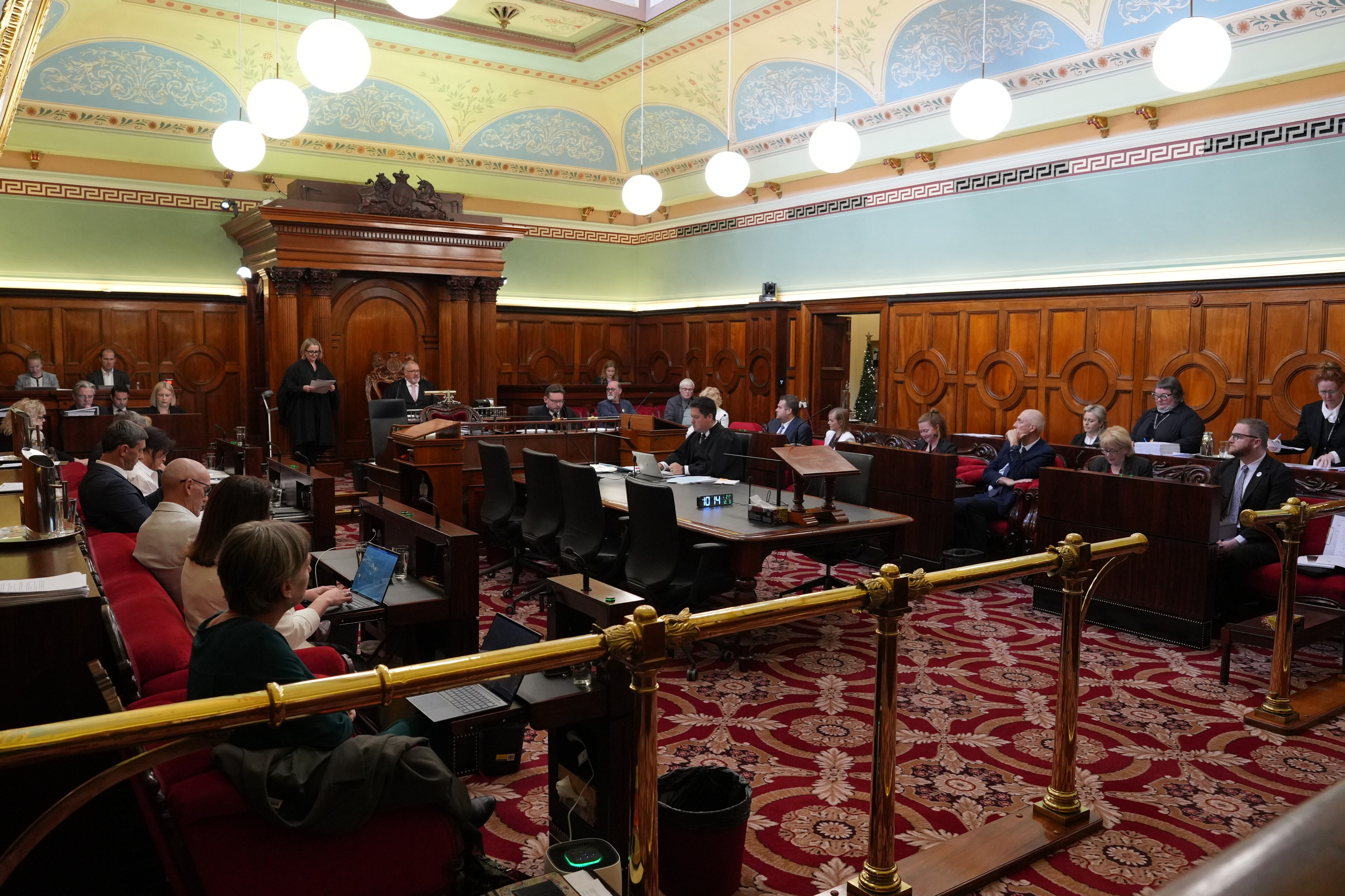 Una toma larga de personas en una habitación ornamentada con paneles de madera y una alfombra roja estampada.