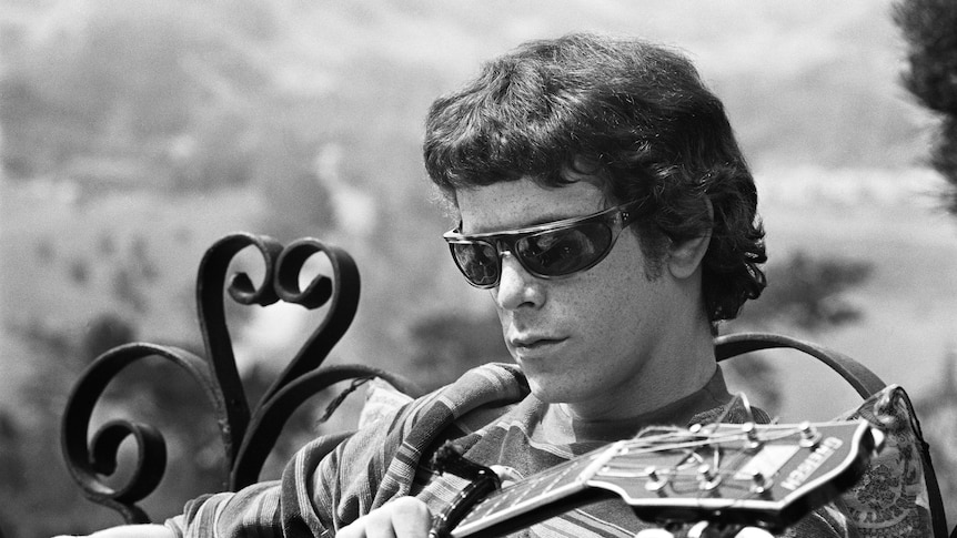 A black and white image of Lou Reed sitting on a bench wearing sunglasses as he plays guitar