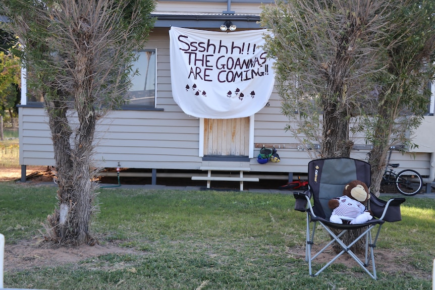 A white house with two trees in front has a big sign hanging from the awning that says "shhhh, the goannas are coming'