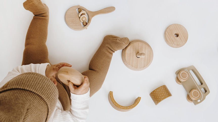 Baby dressed in beige playing with minimalist wooden toys on the floor, viewed from above