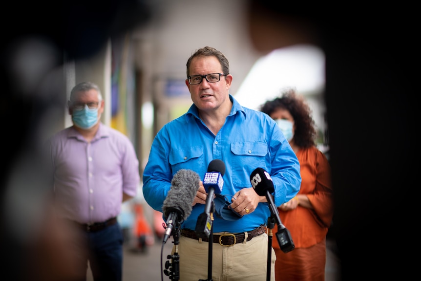 A man in a blue button up shirt standing in front of two microphones at a press conference, flanked by colleagues