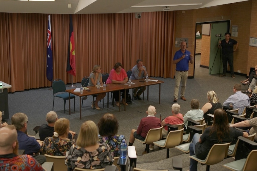 Four people sitting at a table, addressing a crowd of people at a public forum