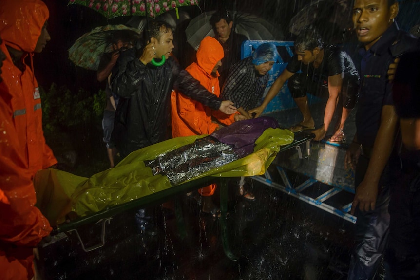 Men, some holding umbrellas, carry a wrapped up body on a stretcher.