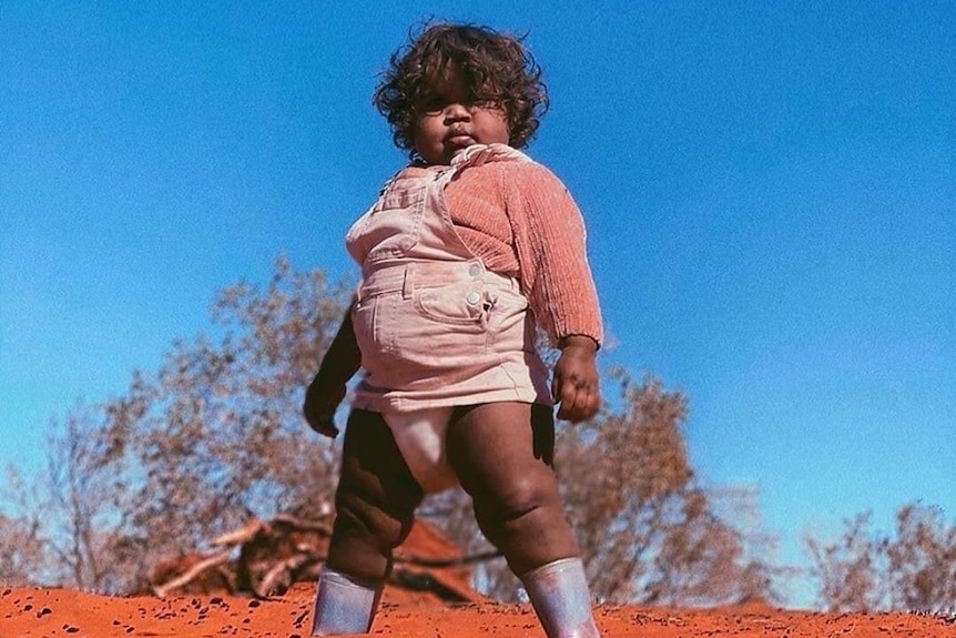 Photo of toddler posing on red sand dune in pink overalls.
