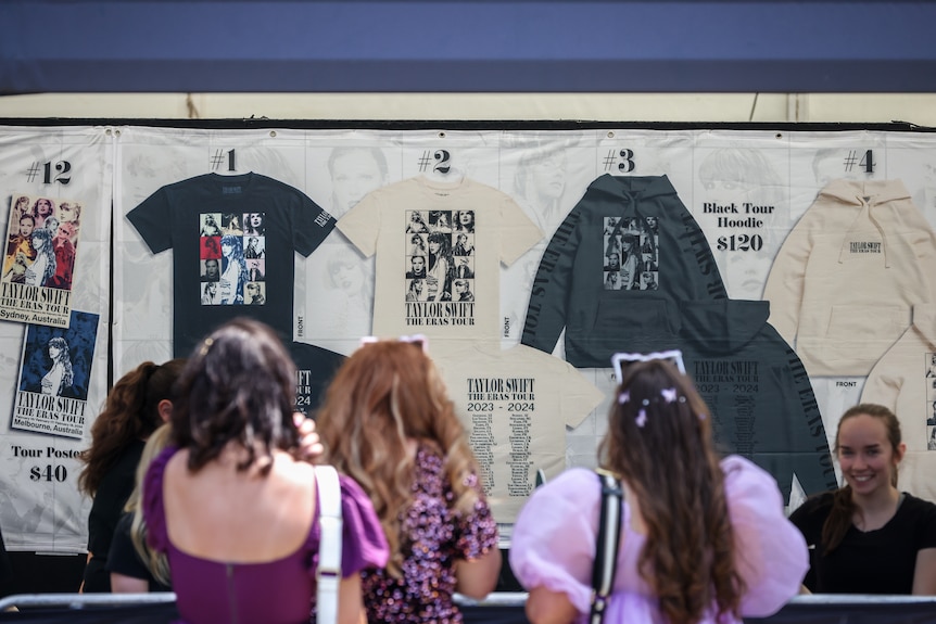 Mujeres jóvenes vestidas de púrpura y destellos se alinean en un puesto de productos donde se venden camisetas de Taylor Swift.
