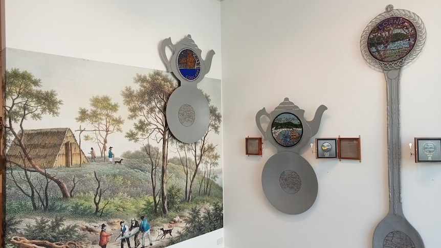 Two giant tea caddies and a giant teaspoon sit on the walls of a gallery, next to wooden boxes containing descriptive text.