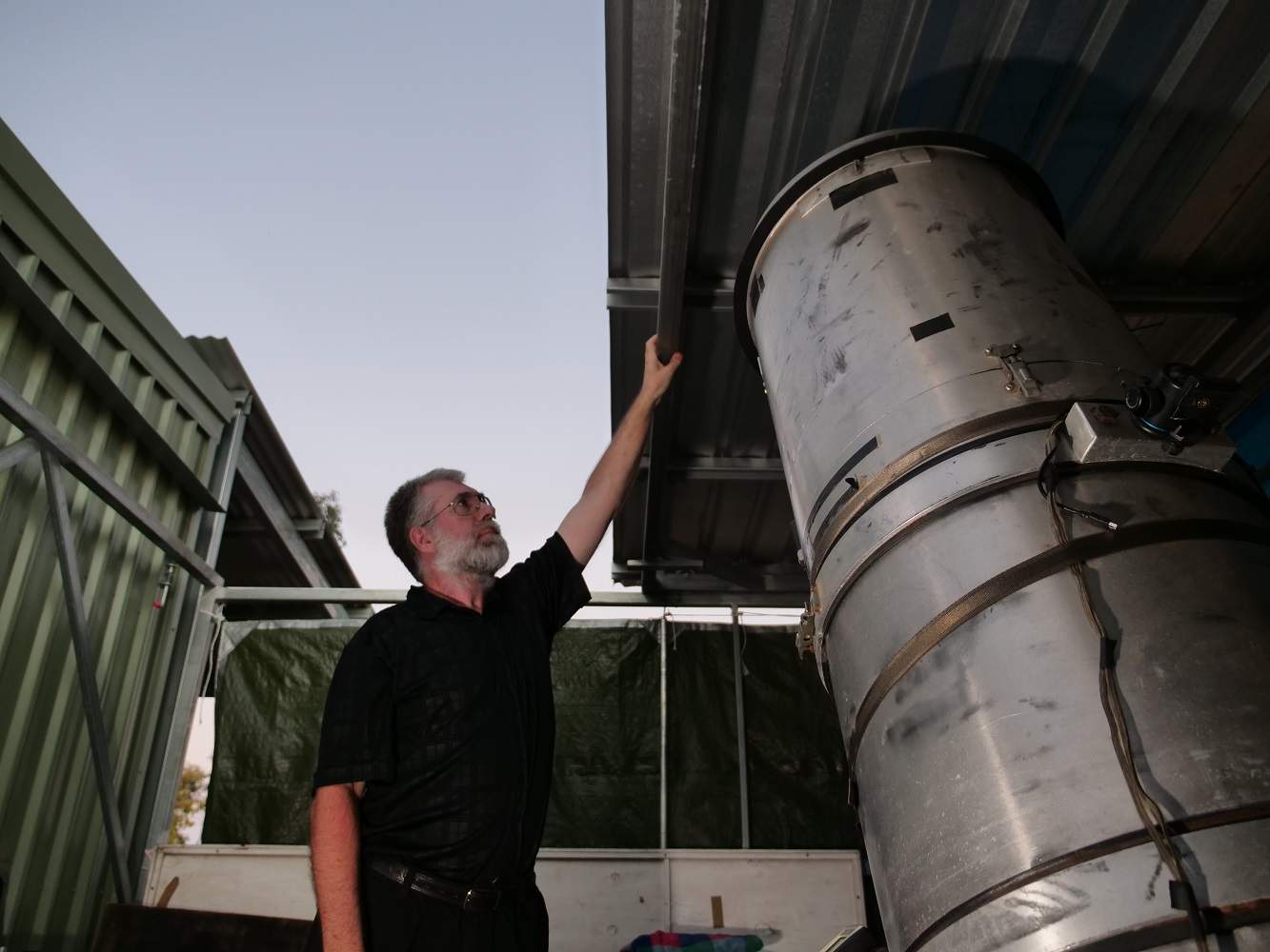 A man with a grey hair stands in a shed and pushes the roof back to the sky at dusk. Inside the shed is a large telescope.