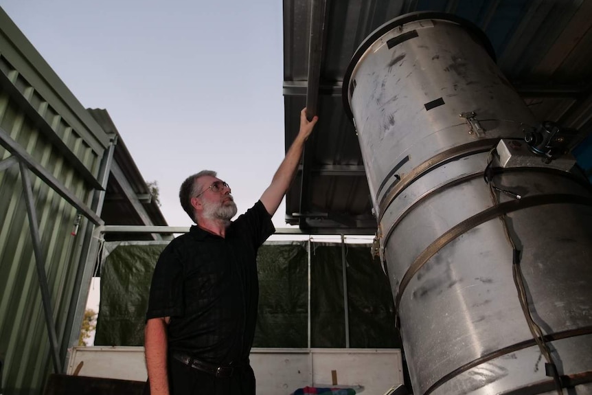 A man with grey hair stands in a shed and pushes the roof back to the sky at dusk. Inside the shed is a large telescope.