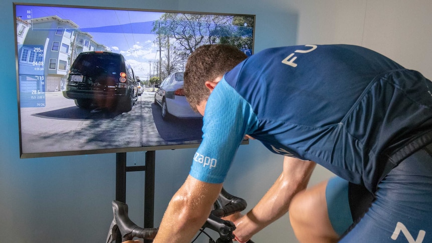 A man is riding a stationary bicycle in front of a screen showing the virtual ride he's taking