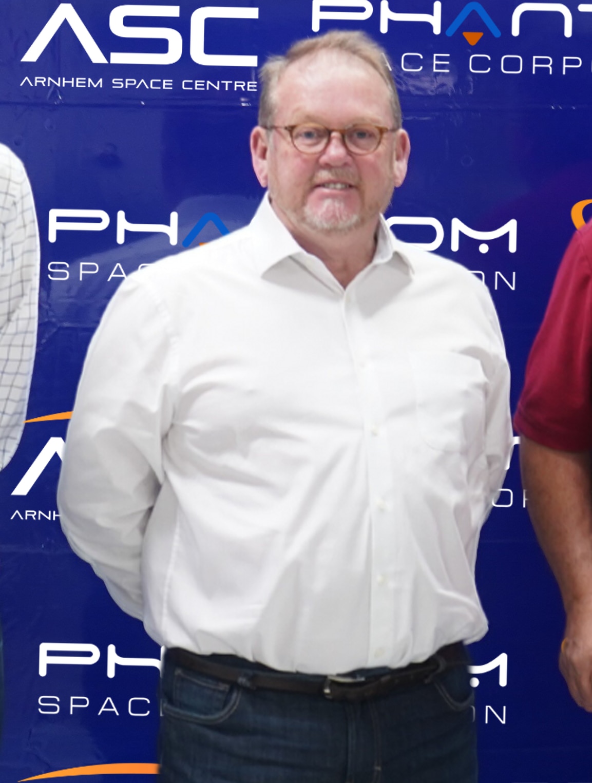 A man wearing glasses and a white dress shirt stands in front of a blue banner