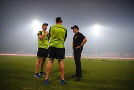 Three men stand together talking on a cricket pitch surrounded by a thick haze.