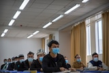 Rows of men sit at desks in a classroom wearing masks. 