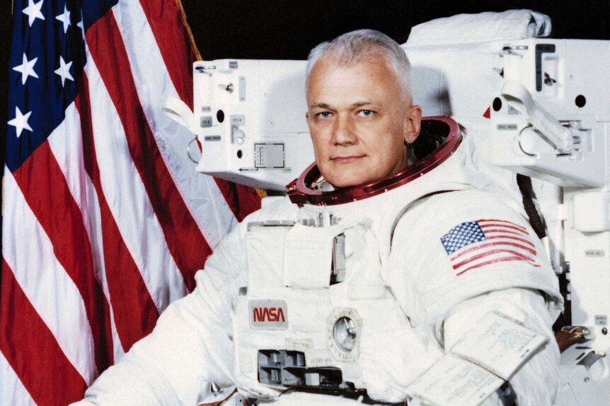 Astronaut Bruce McCandless II poses for the camera wearing his space suit with his helmet off in front of an American flag
