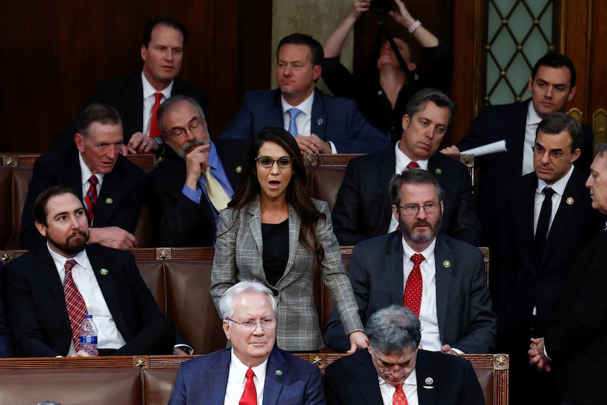 A woman stands in Congress surrounded by men in suits. 