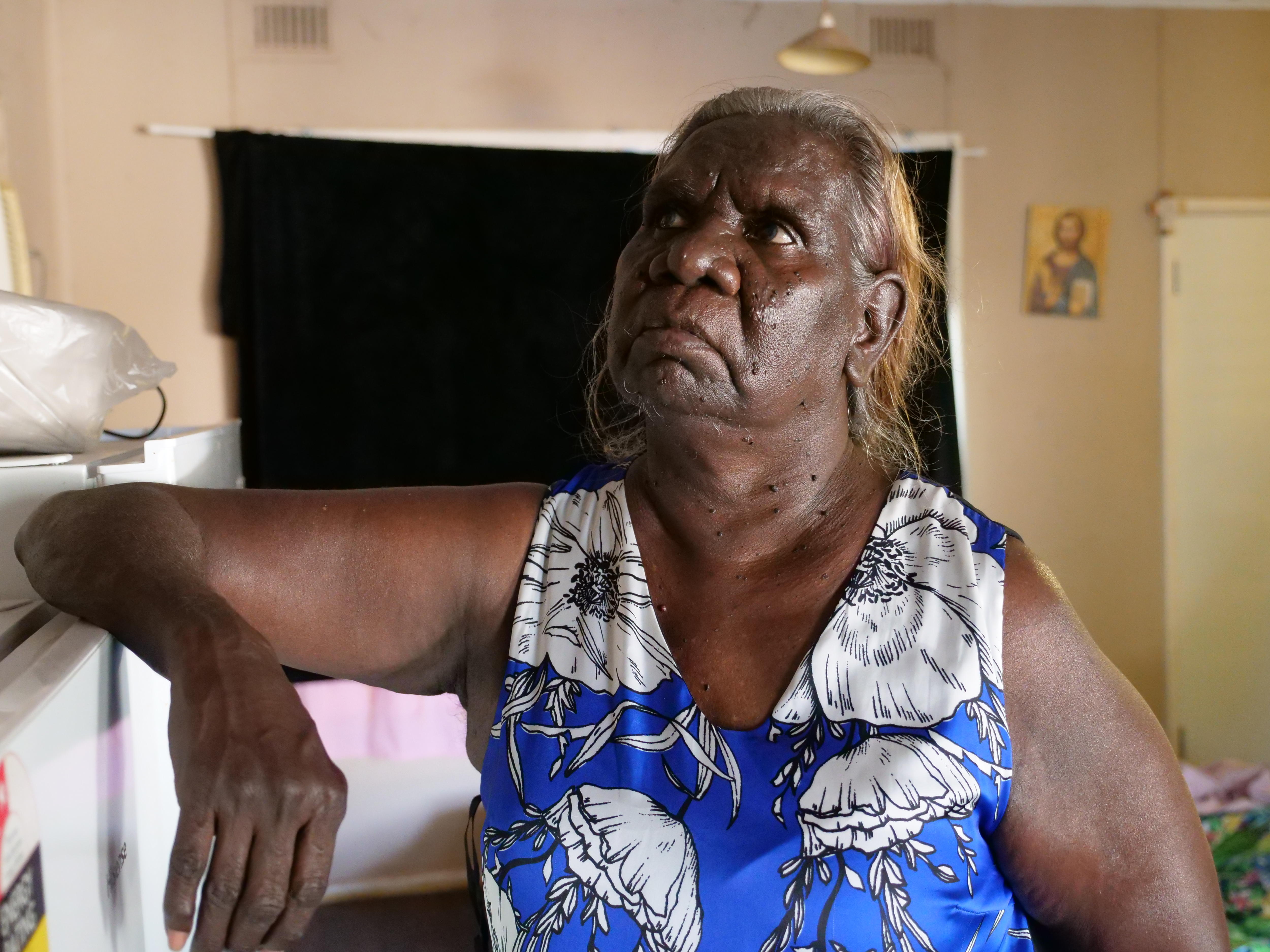 An older Indigenous lady wears a blue dress and looks upwards as she places her elbow on a fridge