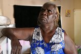 An older Indigenous lady wears a blue dress and looks upwards as she places her elbow on a fridge