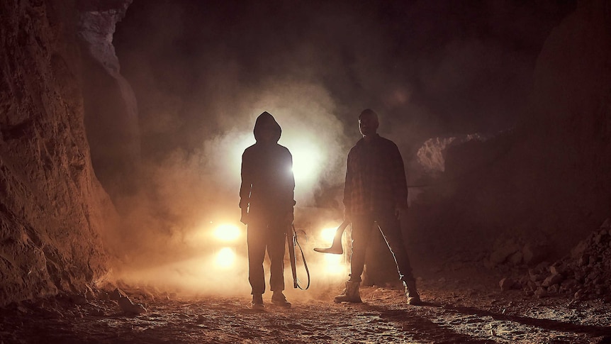 Two people standing together, silhouetted by car headlights. One is holding a boomerang in hand.