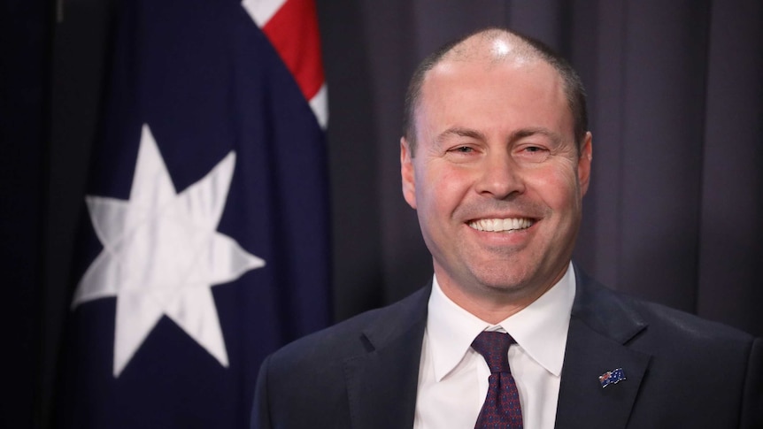 Frydenberg smiles, standing in front of an Australian flag.