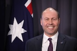 Frydenberg smiles, standing in front of an Australian flag.