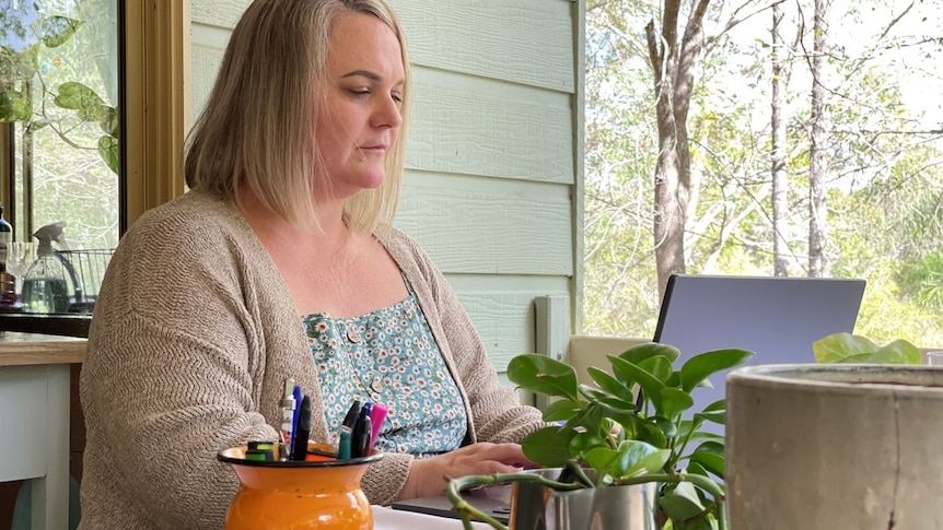 Large middle-aged woman, looks at computer, serious, blonde hair, cardigan, sits in front of weatherboard house, pot plants.