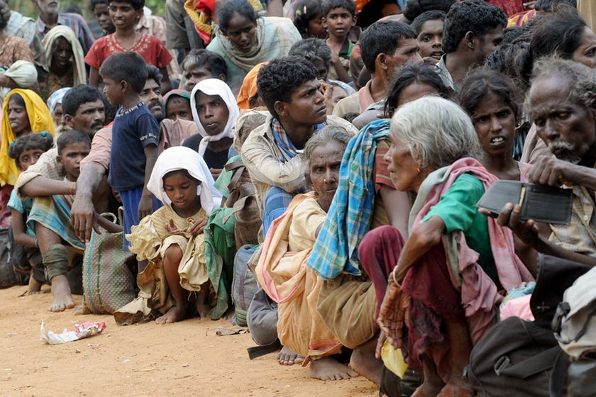 A large group of Tamils crouch together on dirt.
