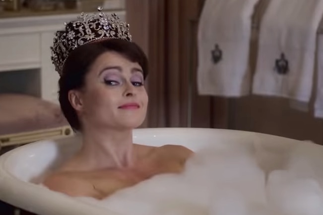 A fair skinned woman with dark hair wears a tiara while sitting in a bubble bath.
