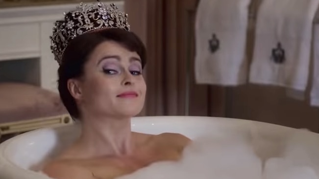 A fair skinned woman with dark hair wears a tiara while sitting in a bubble bath.