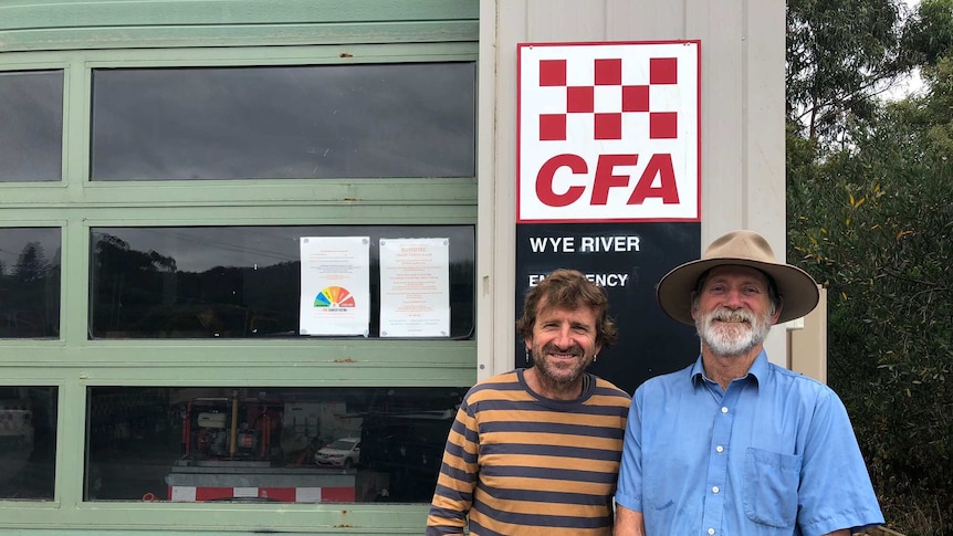 Wye River CFA captain Andrew Hack stands next to former brigade captain Roy Moriarty in front of the station sign.