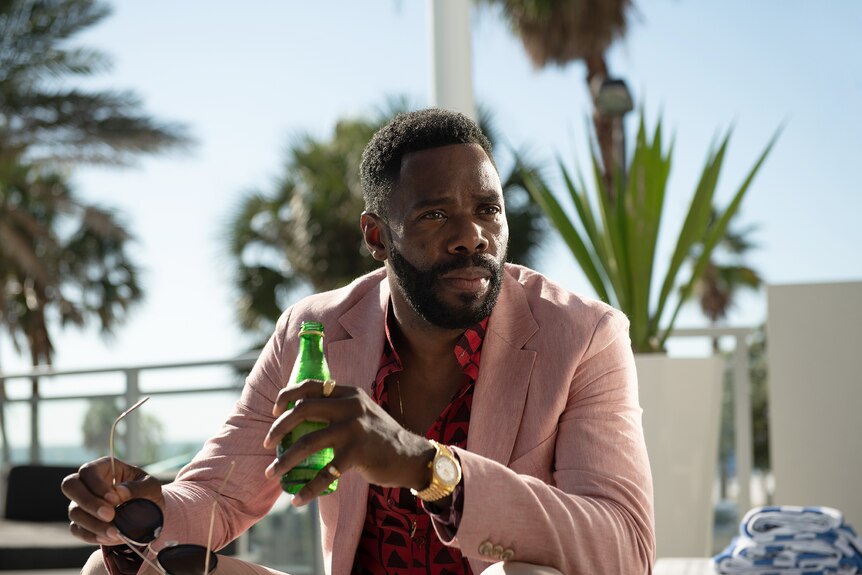 Black man with short hair and beard wears pink blazer over patterned red shirt and sips bottled water in the sun by a pool.
