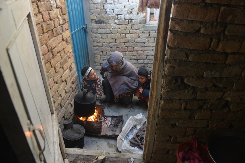 A woman and two young children squat next to an outdoor fire with a pot hanging over it.