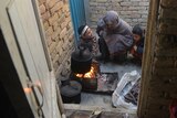 A woman and two young children squat next to an outdoor fire with a pot hanging over it.
