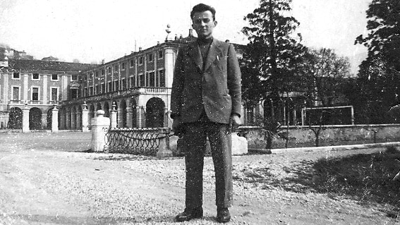 Black and white photo from the 1950s of a young man in suit, standing on a dirt road with mansion-type building behind him.