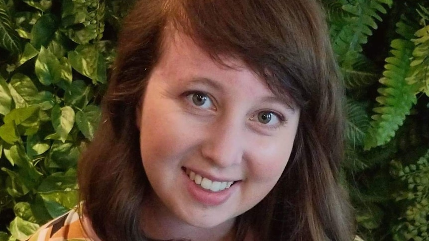 Zoe smiles at the camera in front of a background of plants.