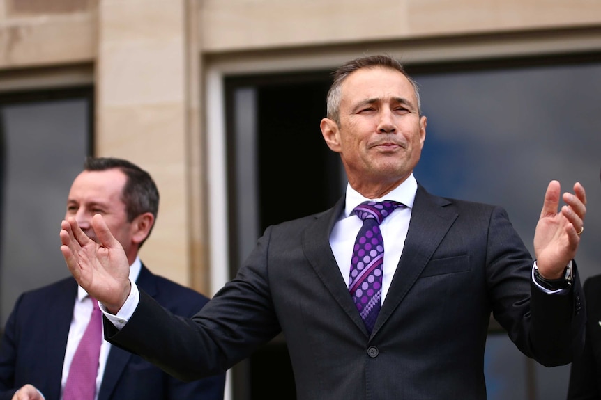Roger Cook stands on the steps of parliament smiling with his arms outstretched. He wears a suit.