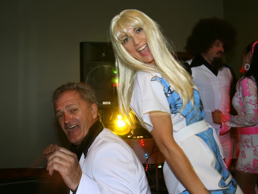 A woman wearing a long blonde wig and a middle-aged man wearing a tuxedo do the boogie at a 70s-themed party.