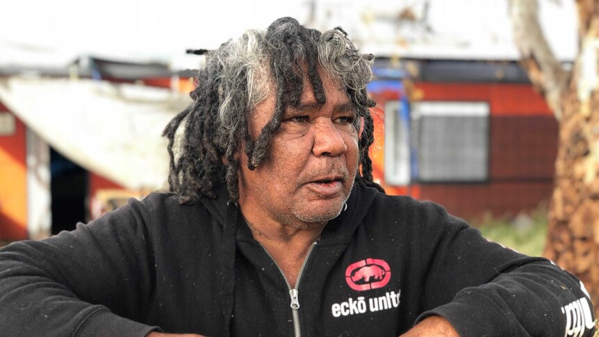 An Aboriginal man leans on a fence with a mug.