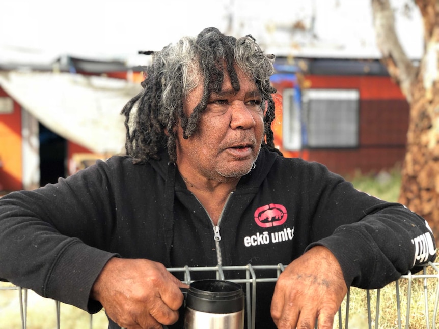 An Aboriginal man leans on a fence with a mug.