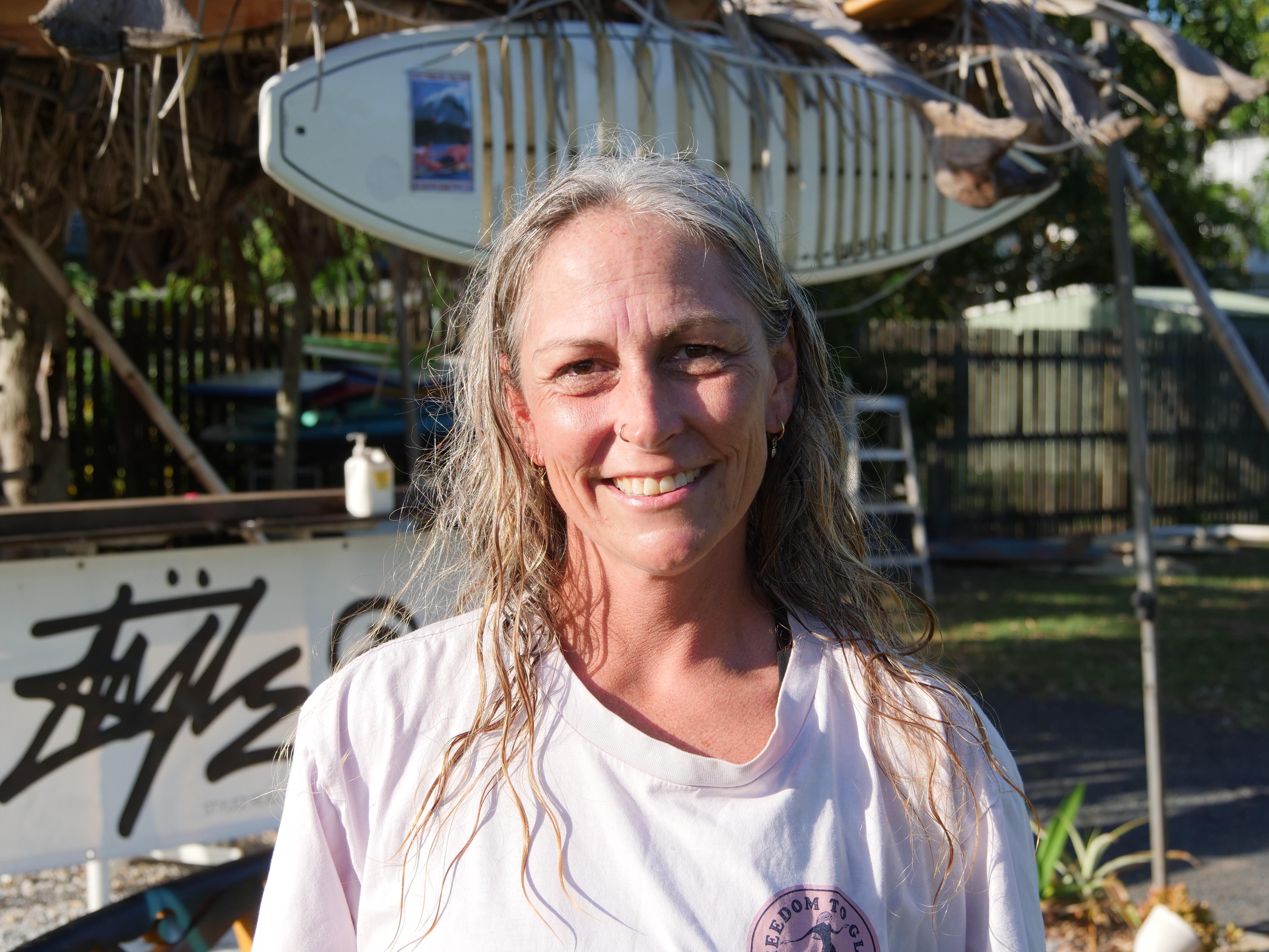 Mujer de pelo largo y claro, con camiseta y sonriendo a la cámara, con una tabla de surf al fondo.