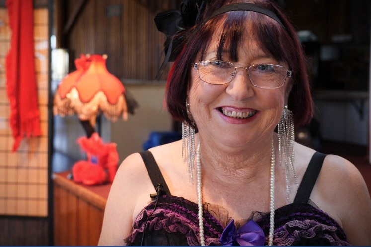 Woman in her 60s with long sparkly earrings and a frilly purple corset smiling widely, lamp and feather boa behind her. 