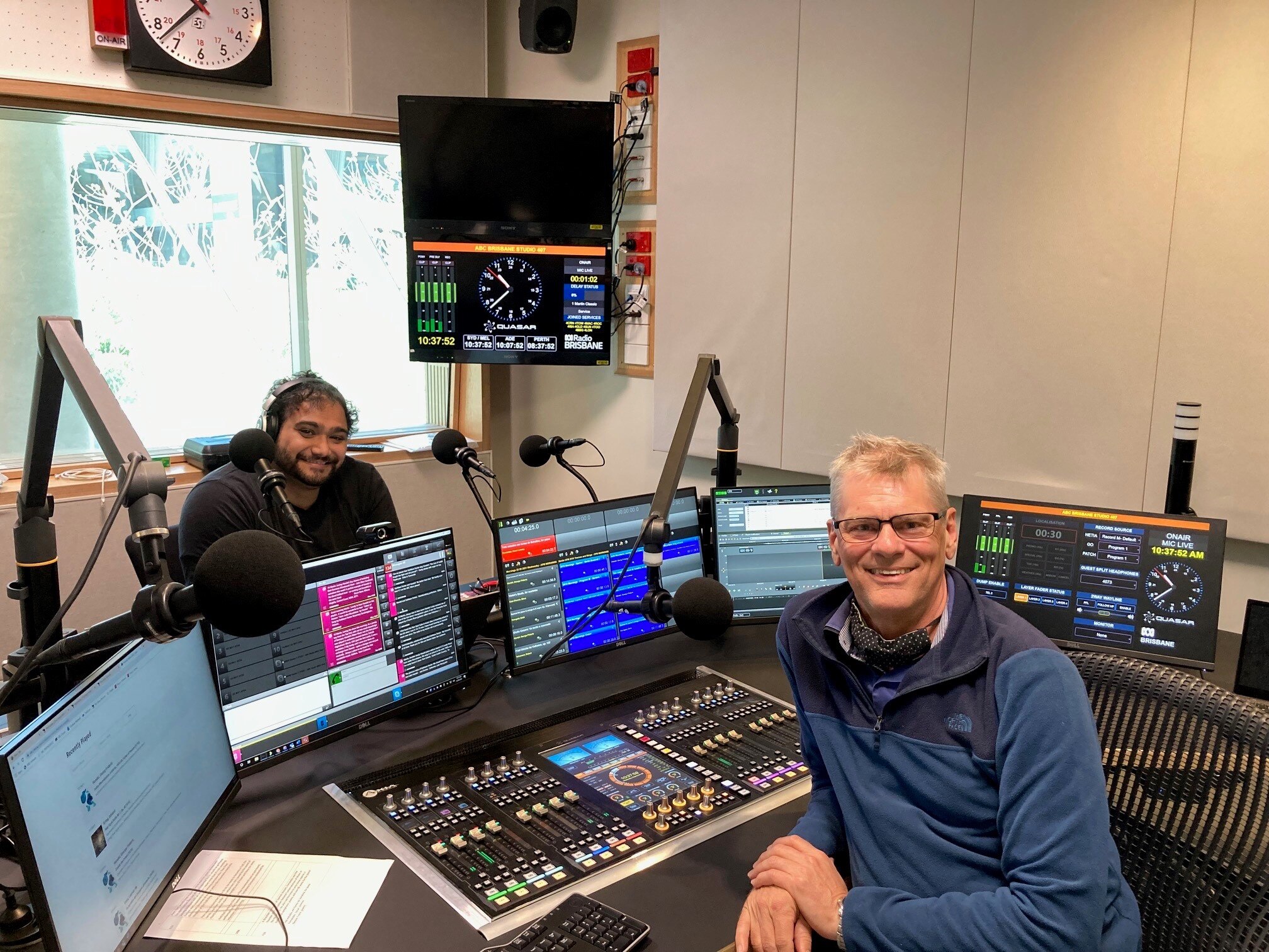 Martin sits at a radio desk with a guest, they both face and smile at the camera.