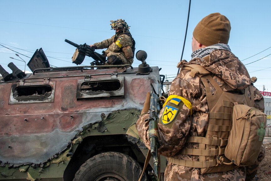 Two men in Ukrainian army uniforms inspect a damaged vehicle.