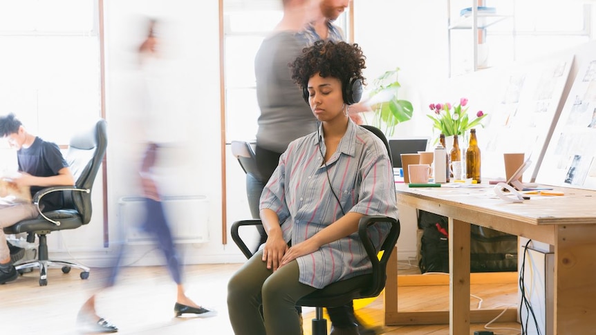 In a room full of people moving and slightly blurred, a woman sits next to a desk with eyes closed and wearing headphones.