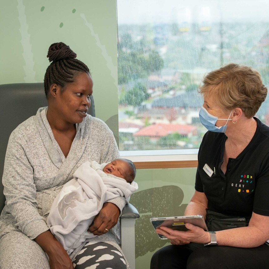 a woman with dark skin holds a sleeping baby looking at an older pale skinned woman who may be a midwife collecting information