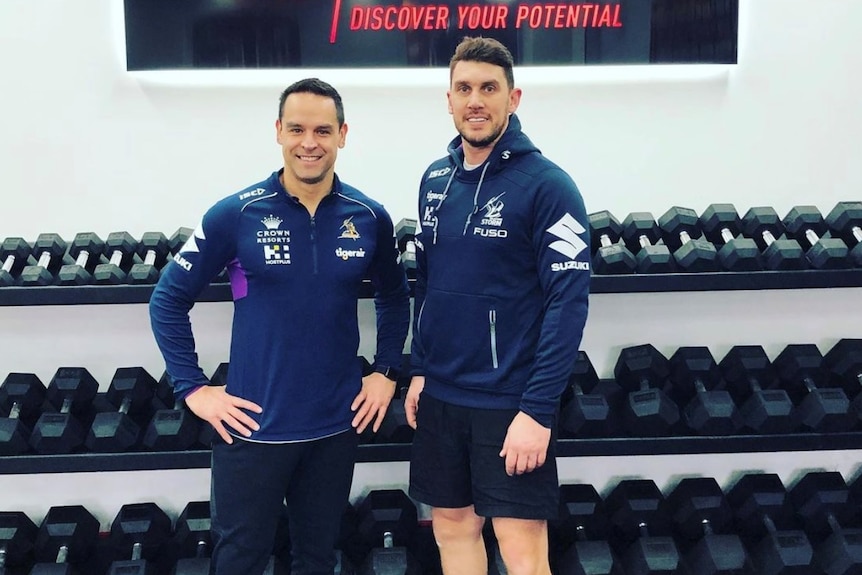 Two men stand in front of racks of dumbbells.