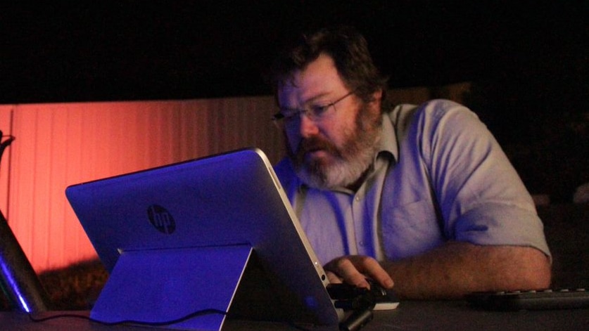 A heavyset middle-aged man, bearded and bespectacled, hunched over a computer.