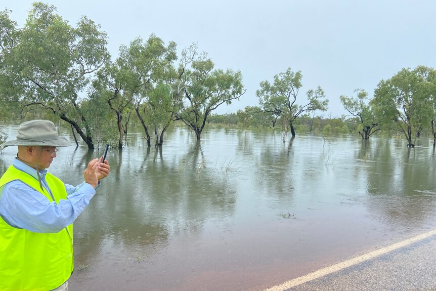 A Main Roads inspector takes a photo of the road on his phone, with flooded trees in the background.