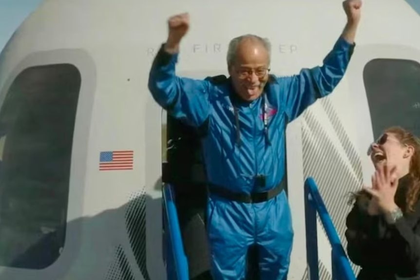 A man in a blue flightsuit emerges from a space capsule with his arms raised.